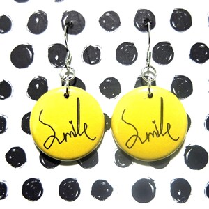 May include: A pair of yellow circular earrings with the word "Smile" in black script. The earrings hang from silver-colored hooks. The background is white with a pattern of black polka dots.