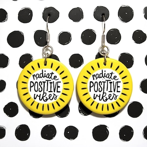 May include: A pair of yellow circular earrings with the words "radiate POSITIVE vibes" in black script. The earrings have a sunburst design around the edges and silver-colored hooks.