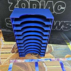 Könnte beinhalten: Ein blaues, mehrstufiges Organizer mit gebogenen Schlitzen. Der Organizer besteht aus Kunststoff und ist für kleine Gegenstände konzipiert. Der Hintergrund enthält den Text "ZODIAC" und "WS".