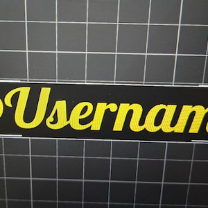 May include: A black rectangular sticker with the text "@Username" in yellow cursive font.