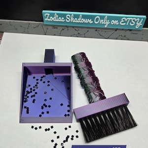 May include: A purple and black 3D printed brush and dustpan set with a black bristle brush and a small dustpan with a black square design. The set is labeled "Aurora Silk".