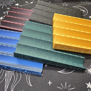 May include: A collection of tiered display stands in various colors, including blue, green, yellow, black, and burgundy. Each stand has a curved front and the text "Ridiculously Magical 2025" engraved on the front.
