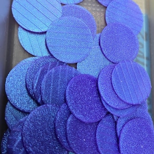 Op de afbeelding: Een close-up van talrijke kleine, cirkelvormige schijven in tinten paars en blauw. Elke schijf heeft een textuuroppervlak en een gestreept patroon. Deze componenten worden waarschijnlijk gebruikt bij het knutselen of als decoratieve elementen.