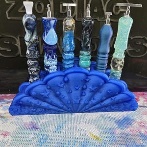 Op de afbeelding: Een blauwe schelpvormige standaard met reliëfsterren bevat zes decoratieve pennen. De pennen hebben gevarieerde ontwerpen, waaronder marmerpatronen, glitters, blauwtinten en metalen accenten. De standaard staat voor een zwart bord met de tekst ZODIAC SHADOWS.