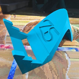 May include: A bright teal-colored stand with the letters "FS" cut out. The stand is designed to hold a craft project. The stand is made of a solid material and has a sturdy base.