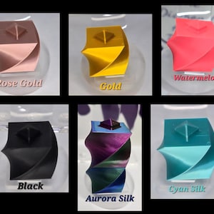May include: A collection of six 3D printed geometric cube objects with a twisted design and a small pyramid on top. The colours include rose gold, gold, watermelon pink, matte black, iridescent aurora silk, and cyan silk, each labelled with its colour name.