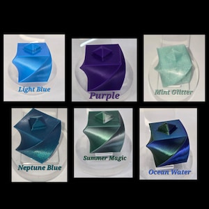 May include: A collection of six 3D-printed geometric sculptures in various colors. The colors include light blue, purple, mint glitter, neptune blue, summer magic, and ocean water. Each sculpture has a square top and a twisted base, with the color name displayed below each.