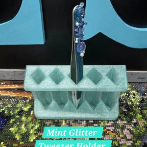 Peut inclure: Un porte-pinces en paillettes vert menthe avec 8 emplacements. Une seule pince à épiler bleue scintillante avec des embellissements en perles et en coquillages est dans le porte-pinces.