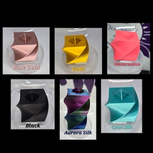 May include: A collection of six 3D-printed geometric sculptures in various colors, including rose gold, gold, watermelon, black, aurora silk, and cyan silk. Each sculpture has a square top and a twisted, curved design, displayed on a clear stand.