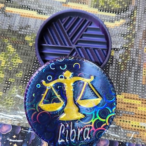 Może przedstawiać: Okrągłe, fioletowe pudełko, wydrukowane w 3D, z wzorem wagi i napisem "Libra" na pokrywce. Pudełko ma holograficzne wykończenie z tęczowymi kolorami.