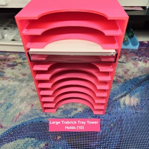 May include: A bright pink storage tower with ten horizontal slots for trays, featuring a semi-circular cutout on each shelf. The unit is labeled Pink Silk and Large Trabrick Tray Tower Holds (10). The structure is designed for organizing small items.