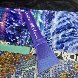 Puede incluir: Una herramienta de pintura de diamantes morada con el texto "Zodiac Shadows" impreso en el mango. La herramienta descansa sobre una pintura de diamantes parcialmente terminada con colores azules y blancos.
