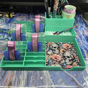 May include: A teal diamond painting organiser with compartments for beads, pens, and accessories. The organiser has a decorative skull and floral design, along with the text "Zodiac Shadows".