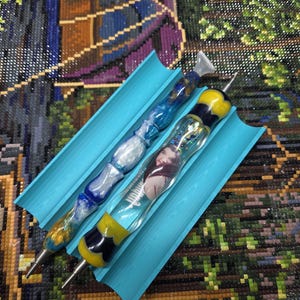 May include: Two diamond painting tools with decorative handles in a blue silicone holder. One tool has a clear handle with a woman's face inside, the other has a blue and white swirl handle.