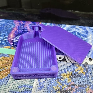 May include: A purple diamond painting tray with a lid. The tray has vertical grooves and the words "Zodiac Shadows" on the side. The lid is rectangular and rests next to the tray. The tray is designed for crafting.