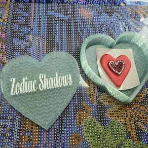 May include: A light teal heart-shaped container with the text "Zodiac Shadows" in white. Inside the container is a red heart with a smaller, embellished heart. The items are on a patterned surface.