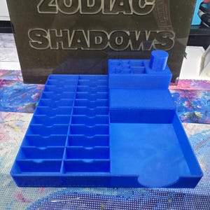 May include: A blue plastic organiser tray featuring a grid of twenty small rectangular slots on the left side, a larger open compartment on the right, and a raised section at the back with various circular and square holes for storage.