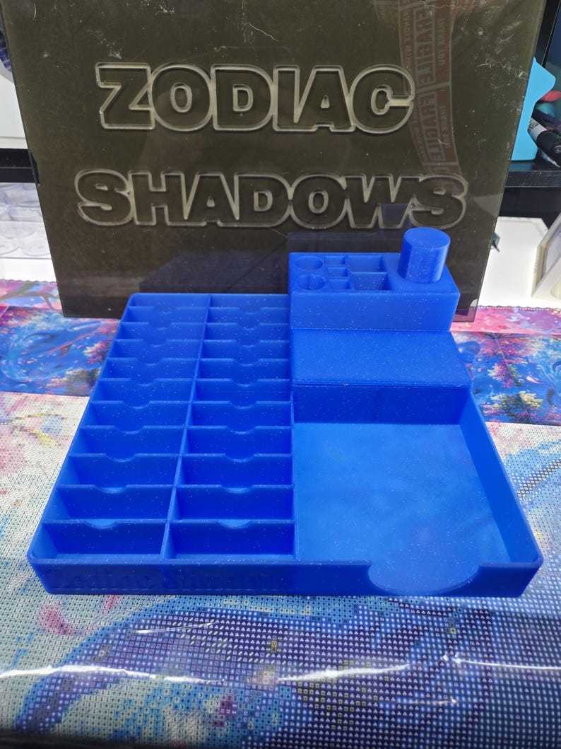 May include: A blue plastic organiser tray featuring a grid of twenty small rectangular slots on the left side, a larger open compartment on the right, and a raised section at the back with various circular and square holes for storage.