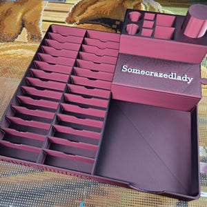 May include: A dark magenta 3D-printed organiser featuring a large grid of twenty-four rectangular slots, a raised section with various small compartments, a cylindrical holder, and a drawer front labelled with the text Somecrazedlady.