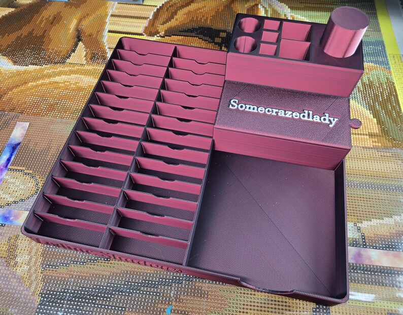 May include: A dark magenta 3D-printed organiser featuring a large grid of twenty-four rectangular slots, a raised section with various small compartments, a cylindrical holder, and a drawer front labelled with the text Somecrazedlady.