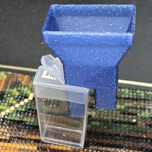 以下が含まれることがあります： 「310」とラベル付けされた透明なプラスチック容器が付いた青いプラスチック製の漏斗。漏斗は、小さなアイテムを容器に注ぐのに役立ちます。