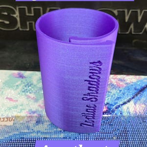 May include: A purple Mega Drill Canvas Roller with a cylindrical shape and an open side. The text Zodiac Shadows is embossed vertically on the side of the roller. The object has a smooth, slightly shimmering finish and is colored in Amethyst.