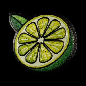 May include: A circular embroidered patch featuring a cross-section of a lime. The design displays a bright yellow-green citrus interior with distinct segments, a thick dark green rind, and a single small green leaf attached to the top edge.
