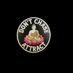 May include: A circular patch with a black background featuring a central illustration of a Buddha figure in yellow robes sitting on a pink lotus flower. The text DON'T CHASE ATTRACT is printed in white capital letters around the central design.