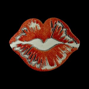 Op de afbeelding: Een decoratieve speld in de vorm van lippen. De lippen zijn oranje-rood gekleurd met een textuurrijke, glinsterende afwerking. Het midden van de lippen is glad en glanzend wit, wat een scherp contrast vormt met de textuurrijke rode randen.