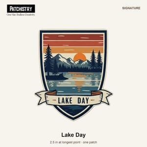 May include: A shield-shaped patch featuring a landscape with a lake, mountains, pine trees, and a sunset. A banner across the bottom displays the text LAKE DAY. The patch measures 6.35 cm at its longest point and has a dark blue border.