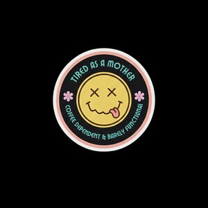 May include: A circular sticker with a black background and a yellow smiley face featuring X-shaped eyes and a tongue sticking out. The text reads TIRED AS A MOTHER and COFFEE DEPENDENT & BARELY FUNCTIONAL, accented by two small pink flowers on the sides.