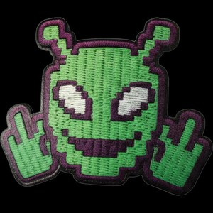Peut inclure: Un écusson brodé représentant une tête d'extraterrestre verte pixelisée avec deux antennes, des yeux blancs et une large bouche violette. L'extraterrestre a deux mains levées de chaque côté, chacune faisant un doigt d'honneur, bordé de fil violet.