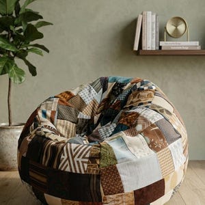 May include: A large, round beanbag chair with a patchwork design. The chair features various fabric patterns in brown, blue, and cream. The beanbag is on a wooden floor, with a plant and shelf in the background.