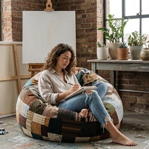 Puede incluir: Una artista dibuja en un cuaderno sentada en un puf de patchwork. La habitación tiene una pared de ladrillo, un caballete de madera con un lienzo en blanco y una mesa con plantas en macetas. Tubos de pintura y pinceles están en el suelo.
