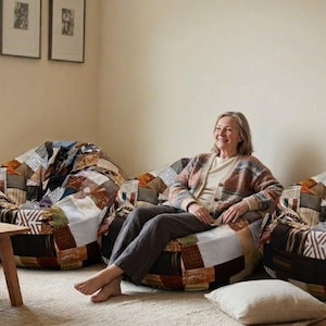 May include: Three large, round bean bag chairs featuring a patchwork design composed of various fabric squares in shades of brown, blue, white, and grey. The chairs have a soft, rounded shape and are arranged in a row on a light-colored floor.
