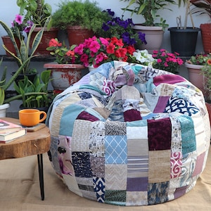 Op de afbeelding: Een patchwork-zitzak met verschillende kleuren en patronen, waaronder blauw, groen, paars en bruin. De zitzak staat op een tapijt voor een muur met planten in potten.
