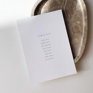 May include: A white card with the text "TABLE SIX" and a list of names rests on a speckled, oval-shaped tray. The tray is a light brown color with darker brown veining. The card is angled, and the background is white.