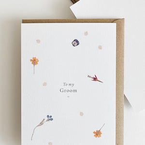 May include: A white card with the text "To my Groom" and a few pressed flowers. The card is surrounded by a brown envelope.