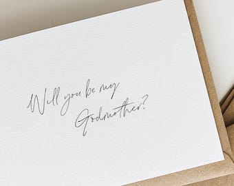 May include: A white card with the handwritten text "Will you be my Godmother?" is displayed on a brown envelope. The card and envelope are on a white surface. The text is in a delicate, cursive font.