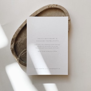 May include: A white wedding invitation card with the names "ERICA MATTHEWS & GRAHAM THOMLINSON" printed in a minimalist font. The card includes event details and is placed on a textured, oval-shaped tray.