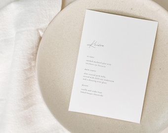 May include: A white rectangular menu card with black text titled Keiron, listing courses including smoked mackerel pate, slow roasted pork belly, and vanilla and tonka bean baked basque cheesecake, placed on a beige ceramic plate.