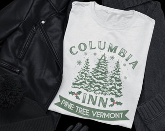 May include: A white t-shirt with the text "COLUMBIA INN PINE TREE, VERMONT" in green, with a graphic of pine trees. The shirt is surrounded by a black leather jacket, black beanie, black gloves, gray jeans, and a gray sweater.