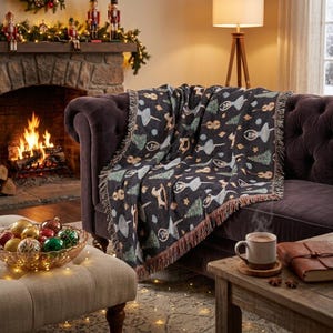 May include: A dark gray woven throw blanket featuring a repeating pattern of ballet dancers, Christmas trees, and stars. The blanket has a fringed edge and is draped over a dark brown tufted sofa in a room with a fireplace and holiday decor.