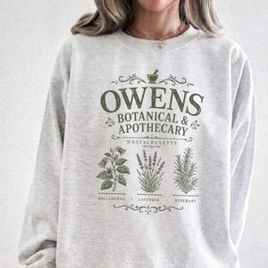 Könnte beinhalten: Ein hellgraues Sweatshirt mit Rundhalsausschnitt und dunkelgrünem Text, der OWENS BOTANICAL & APOTHECARY MASSACHUSETTS lautet. Unter dem Text befinden sich Illustrationen von Tollkirsche, Lavendel und Rosmarin mit den Namen darunter.