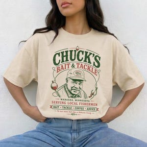 Könnte beinhalten: Ein beiges Kurzarm-T-Shirt mit einer Grafik eines Mannes mit Kappe und dem Text "CHUCK'S BAIT & TACKLE WABASHA, MINNESOTA SERVING LOCAL FISHERMEN BAIT TACKLE COFFEE ADVICE The best damn bait shop in Wabasha County".
