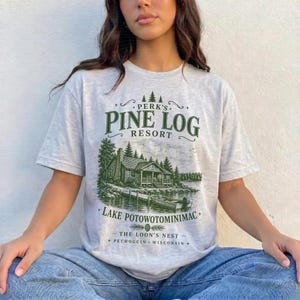 May include: A light gray short-sleeve t-shirt featuring a dark green graphic of a cabin by a lake. The text reads: PERK'S PINE LOG RESORT, LAKE POTOWOTOMINIMAC, THE LOON'S NEST, PECHOGGIN WISCONSIN. The design includes a small boat and pine trees.