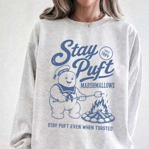 May include: A light gray crewneck sweatshirt featuring a blue graphic of the Stay Puft Marshmallow Man roasting a marshmallow over a campfire. The text reads Stay Puft, Since 1984, Marshmallows, and Stay Puft Even When Toasted in a vintage-style font.