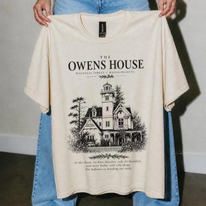以下が含まれることがあります： クリーム色の半袖Tシャツで、ビクトリア様式の家の黒いインク風イラストが描かれています。テキストには「THE OWENS HOUSE MAGNOLIA STREET MASSACHUSETTS」および「In this house, we have chocolate cake for breakfast, and never bother with silly things like bedtimes or brushing our teeth.」と記されています。