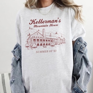 Könnte beinhalten: Ein hellgraues T-Shirt mit Rundhalsausschnitt und einer kastanienbraunen Strichzeichnung eines großen Gebäudes, darunter der Text Kellerman's Mountain House und SUMMER OF '63. Das T-Shirt wird unter einer blauen Jeansjacke mit abgenutzten Ärmeln getragen.