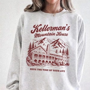 Op de afbeelding: Een lichtgrijs sweatshirt met ronde hals en een donkerrode grafische print. Het ontwerp toont een groot gebouw met bergen en dennenbomen, met de tekst Kellerman's Mountain House erboven en HAVE THE TIME OF YOUR LIFE eronder gedrukt.